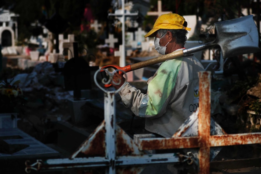 A man wearing a face mask and cap carries a shovel over his shoulder, in a graveyard