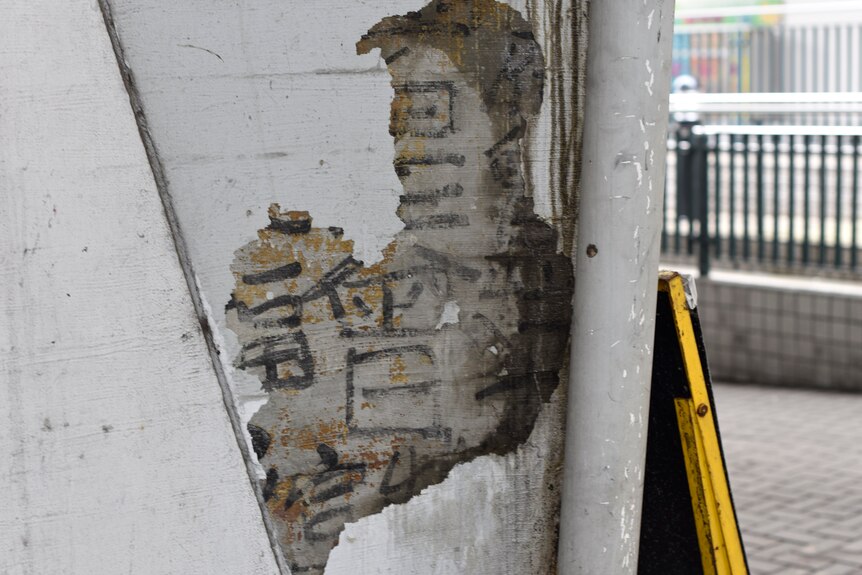 A carpark wall with grey paint chipped away to reveal black calligraphy characters.