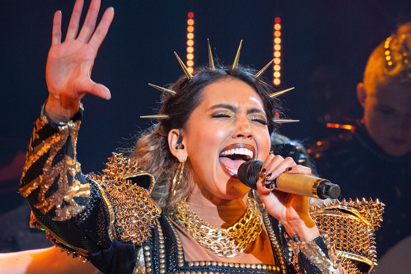 Performer wearing gold spiked headband and gold-studded jacket, holding mic, eyes closed and right hand raised in air, singing.