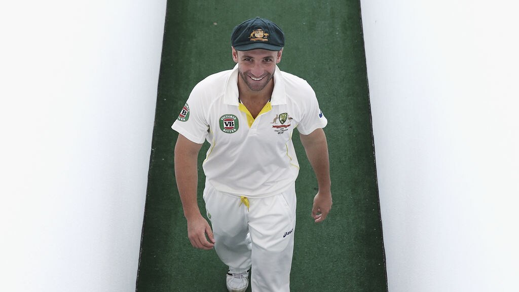 Australian Test cricketer wearing a baggy green walking along a green pathway and looking up smiling at the camera.