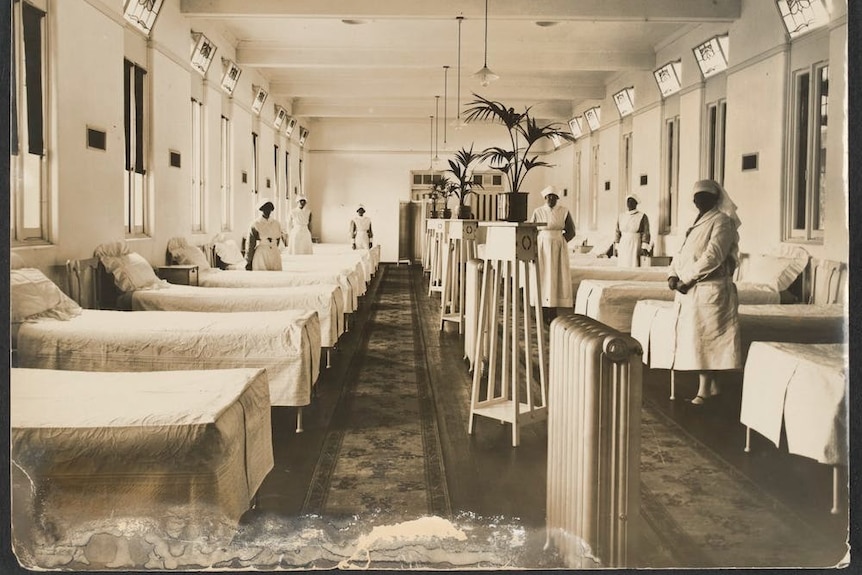 A black and white image of a 1920s hospital ward, with nurses standing around the room.