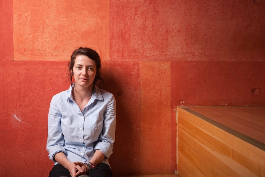 An Aboriginal woman in her 30s wearing a shirt and sitting in front of an orange background