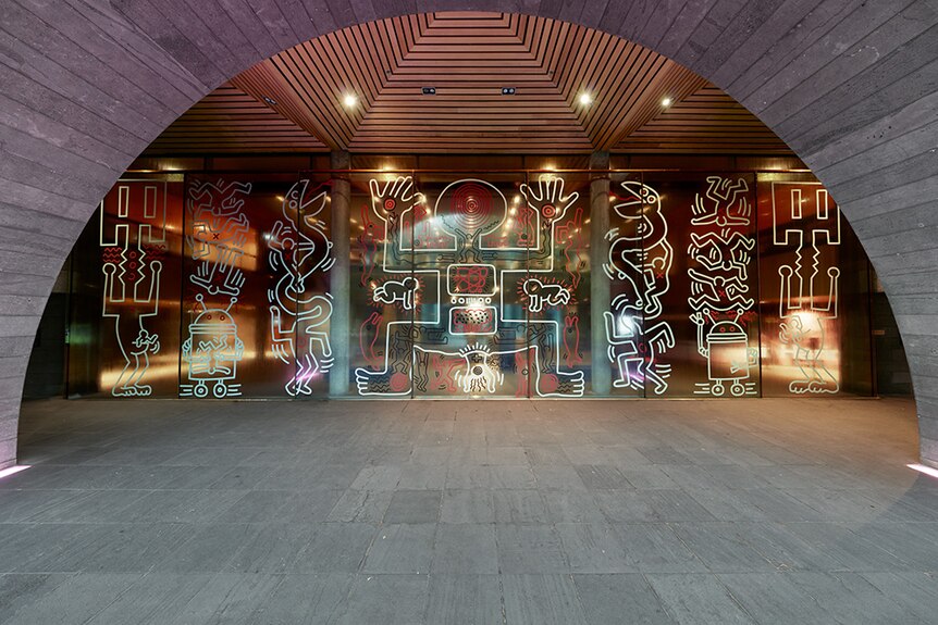 Bold and graphic outlines of shapes and human and imagined creatures on glass wall at concrete and wood museum entrance.