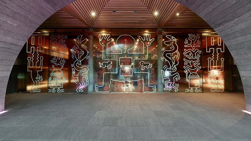 Bold and graphic outlines of shapes and human and imagined creatures on glass wall at concrete and wood museum entrance.