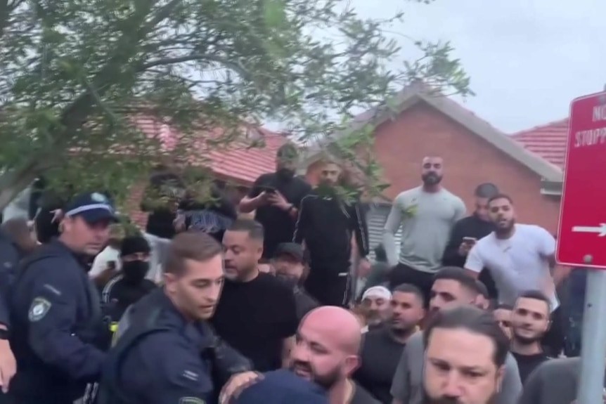 Dozens of people standing on the side of a road with police officers trying to hold them back