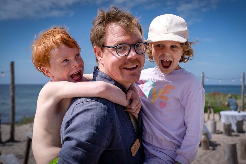 A middle-aged man in glasses carries his two young children near a beach.