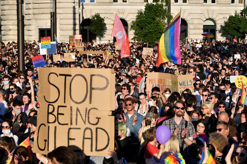 A crowd of LGBT activists in Budapest, one holding a large cardboard sign saying "stop being Gilead".