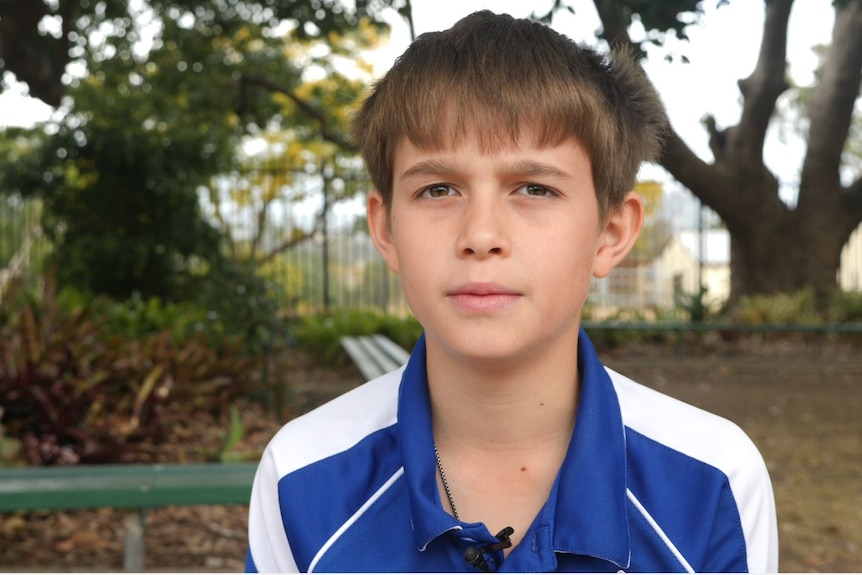 Young boy in a blue and white school sports shirt, a tree behind him and a deep soft stare