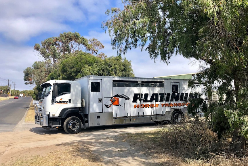 A cab and large trailer with 'horse transport' written on the side drives out the dirt driveway of a regional property.