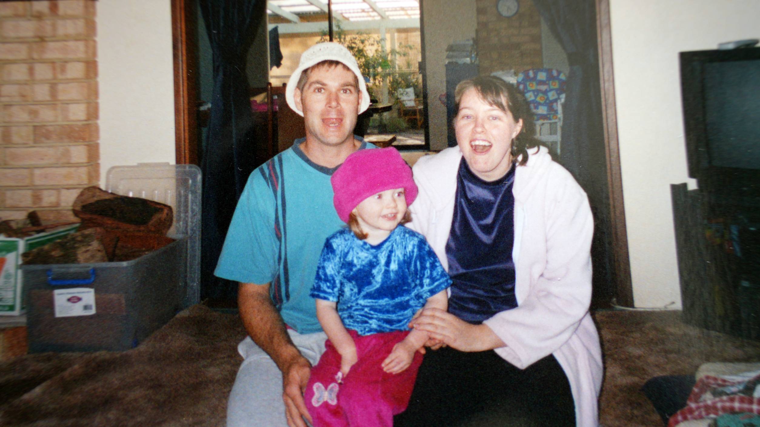 A man with a white hat on smiling, sitting next to a small girl with a pink beanie next to her mother in a white cardigan.