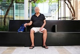 Older white male named Len James wearing black polo shirt, cream shorts and glasses sits on a public bench in Perth city.