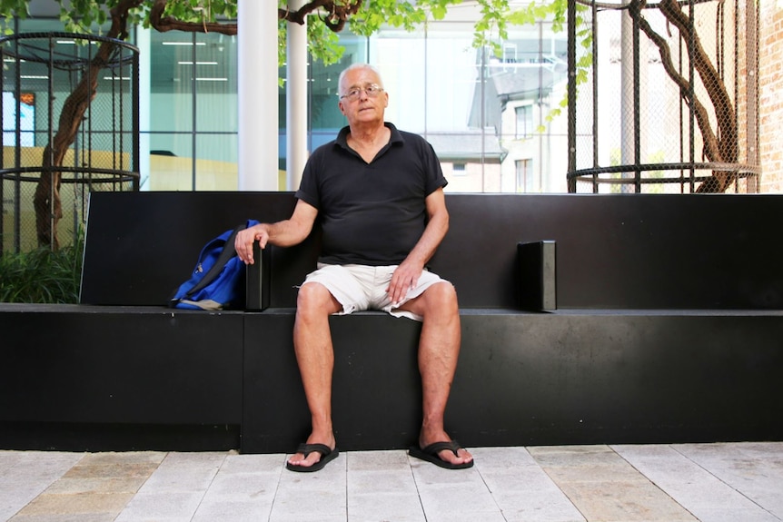 Older white male wearing black polo shirt, cream shorts and glasses sits on a public bench in Perth city.