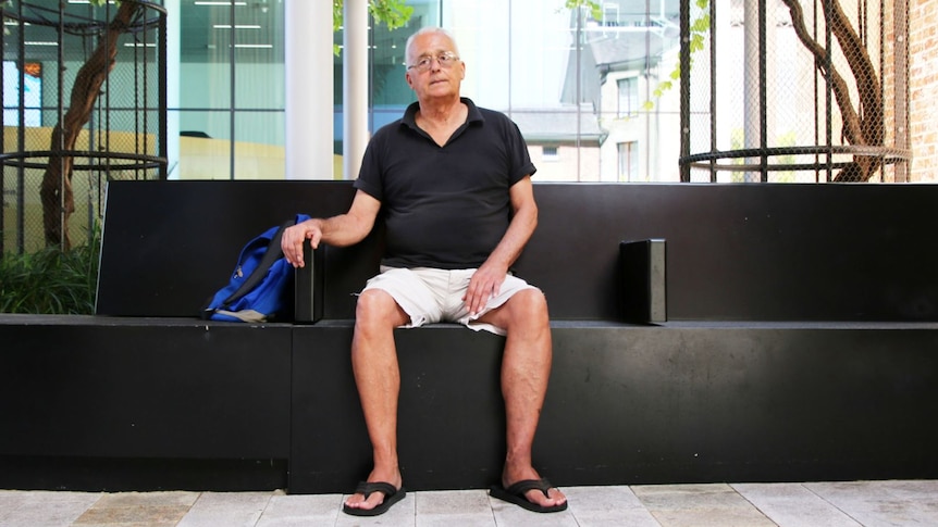 Older white male named Len James wearing black polo shirt, cream shorts and glasses sits on a public bench in Perth city.