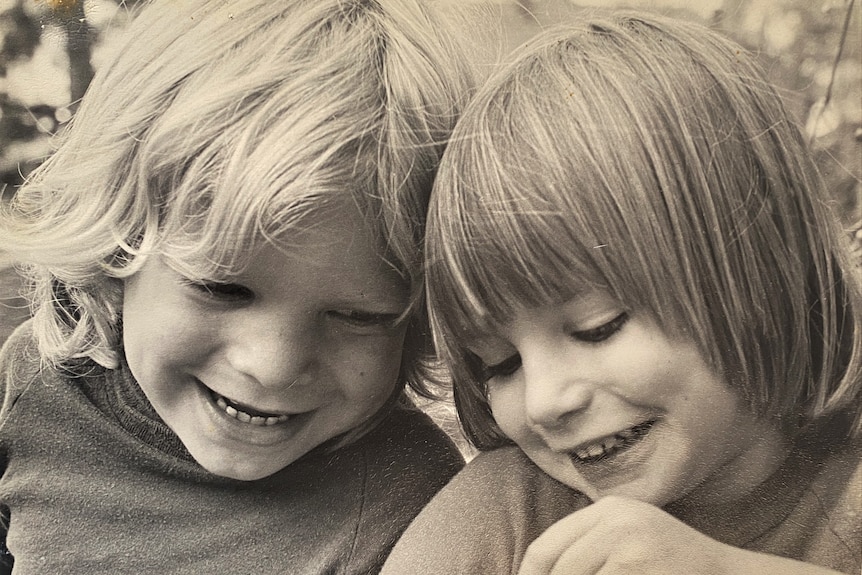 Black and white photograph of two little kids, a boy and a girl. Both are smiling and looking downward