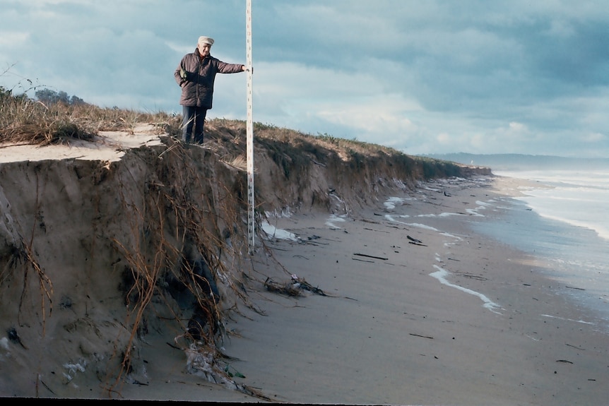 A man stands on top of a sand dune cliff beside the beach, using a long white rod to measure its height.