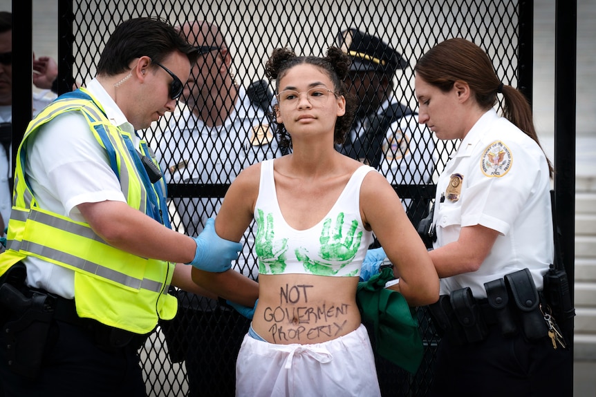 A woman with "Not Government Property" written on her torso is handcuffed by cops 