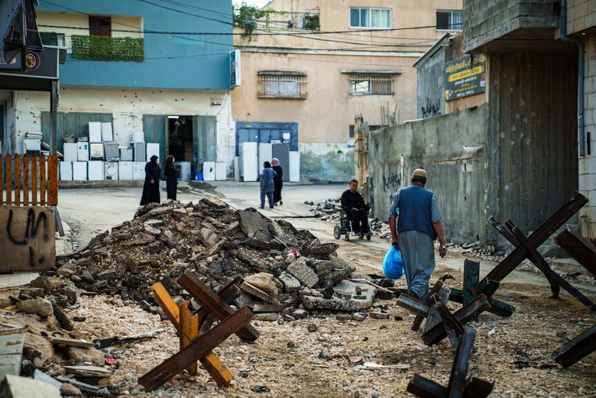 A pile of rubble sits in a road surrounded by people and steel roadblocks.