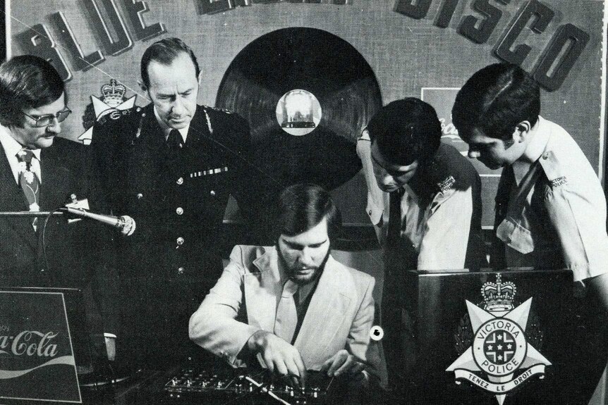 Four men, three in police uniform, watch as another man works a DJ desk. (Black and white)