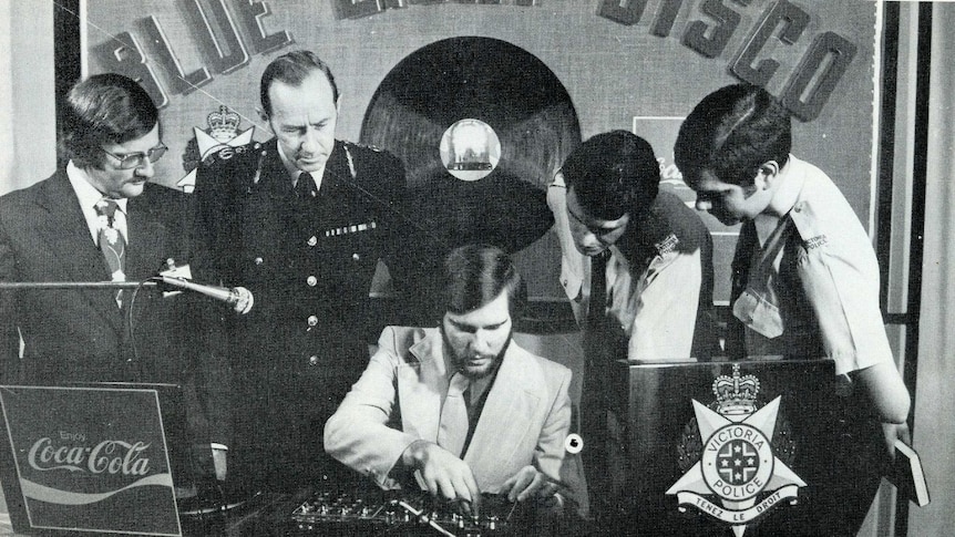Four men, three in police uniform, watch as another man works a DJ desk (black and white photo)