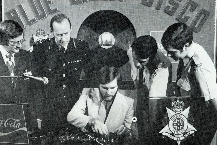 Four men, three in police uniform, watch as another man works a DJ desk. (Black and white)