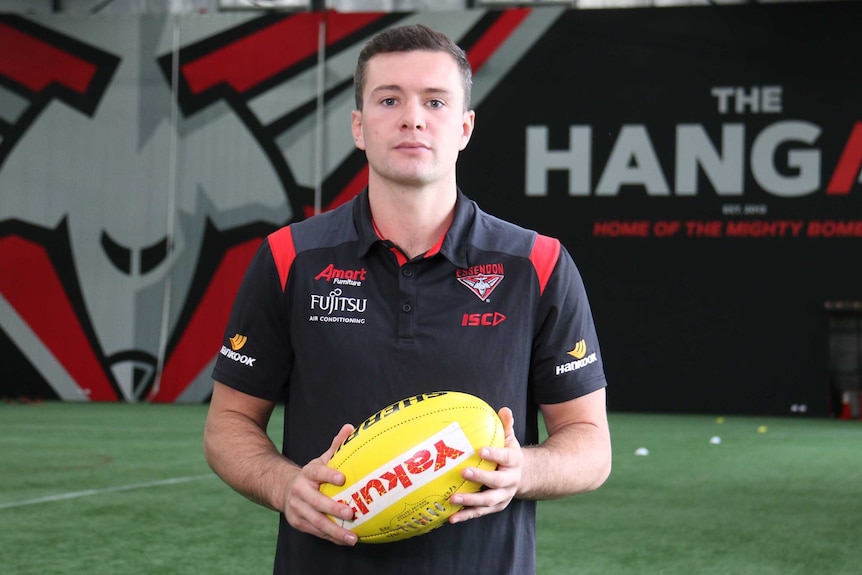 Conor McKenna stands holding a football before a wall with displaying the Essendon logo