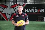 Conor McKenna stands holding a football before a wall with displaying the Essendon logo
