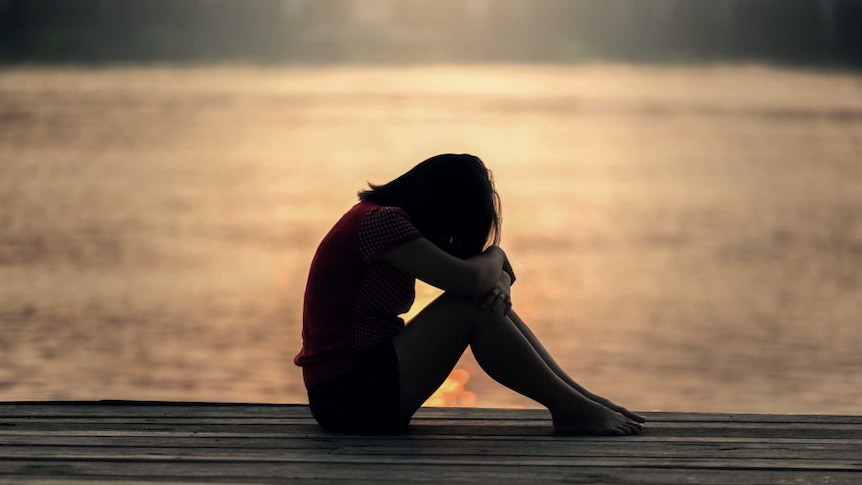 A woman is pictured in silhouette sitting on a jetty with her head on her arms, hugging her knees