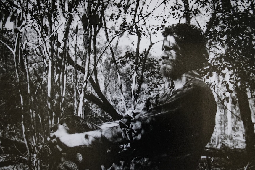 Black and white photo of bearded man in dappled light in coastal forest with an intense expression.