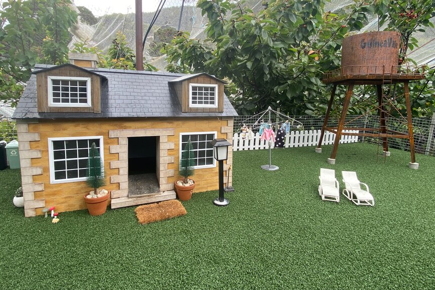A model house on fake grass with white deckchairs outside