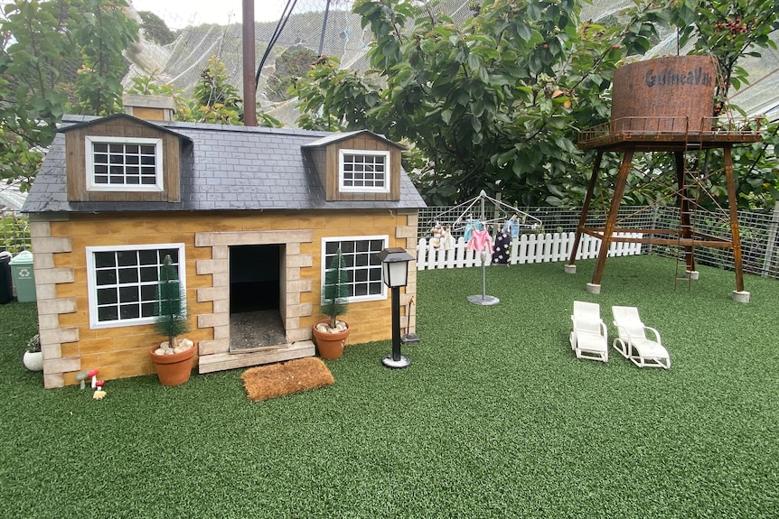 A model house on fake grass with white deckchairs outside
