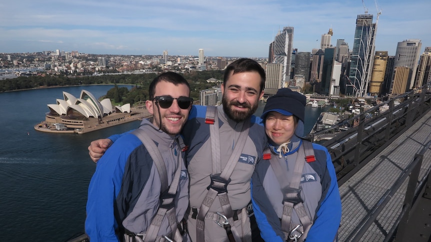 Two men and a woman smile as they have their photo taken on top the Sydney Harbour Bridge.