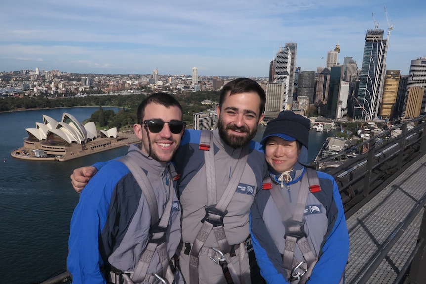 Two men and a woman smile as they have their photo taken on top the Sydney Harbour Bridge.