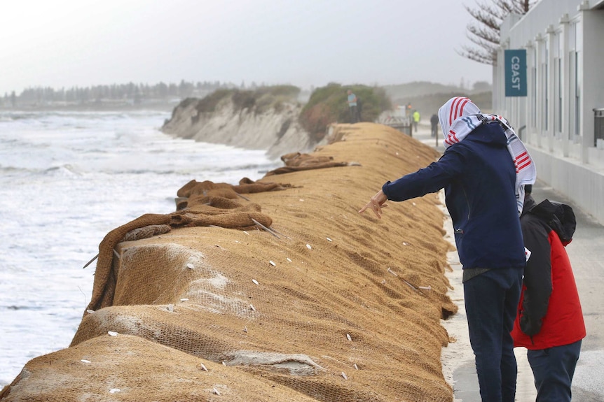 People look on and point at a badly eroded stretch of beach being reinforced by hessian material.