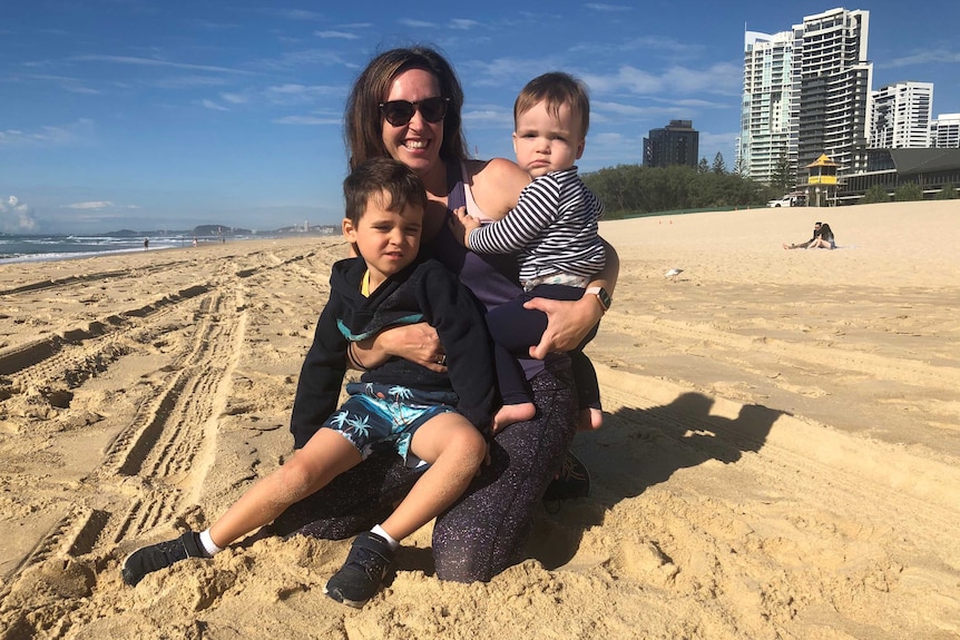 A mother smiles while hugging her two small suns on a sunny stretch of beach.