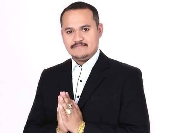 A man wearing a suit holds his hands in prayer position. He wears gold jewellery