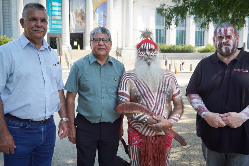 Four Indigenous men, one in traditional dress, stand side by side 