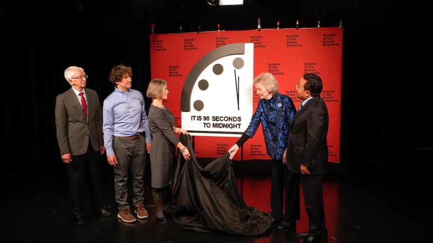 five people standing on black stage, two women removing black cloth from image of a clock