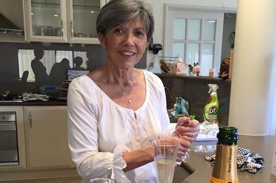 Doreen Collyer stands at a kitchen bench with a bottle of champagne and two glasses in front of her.