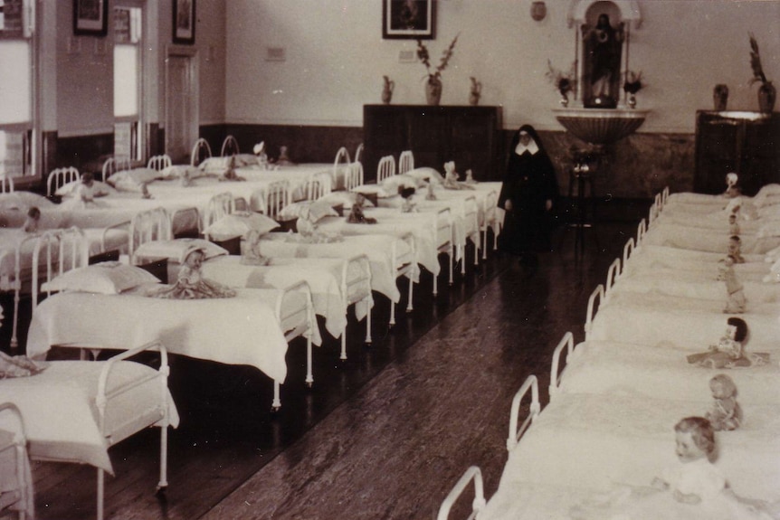 A black and white image of rows of beds with small children in a dormitory and a nun
