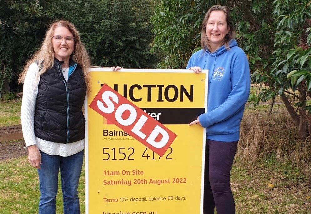 Two women smile as they stand either side of an auction sign with a sold sticker on it.