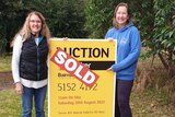 Two women smile as they stand either side of an auction sign with a sold sticker on it.