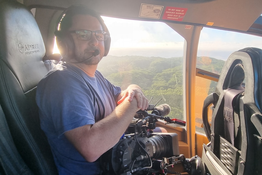 Man holding a camera inside a chopper cabin with green landscape visible through side window.