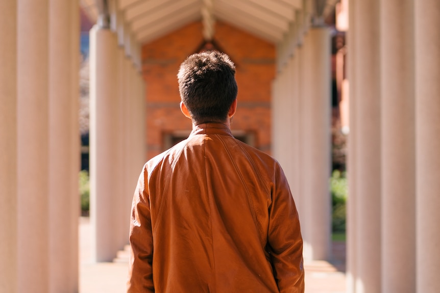 A photo of the back of a man wearing a brown jacket standing in a corridor lined by columns.