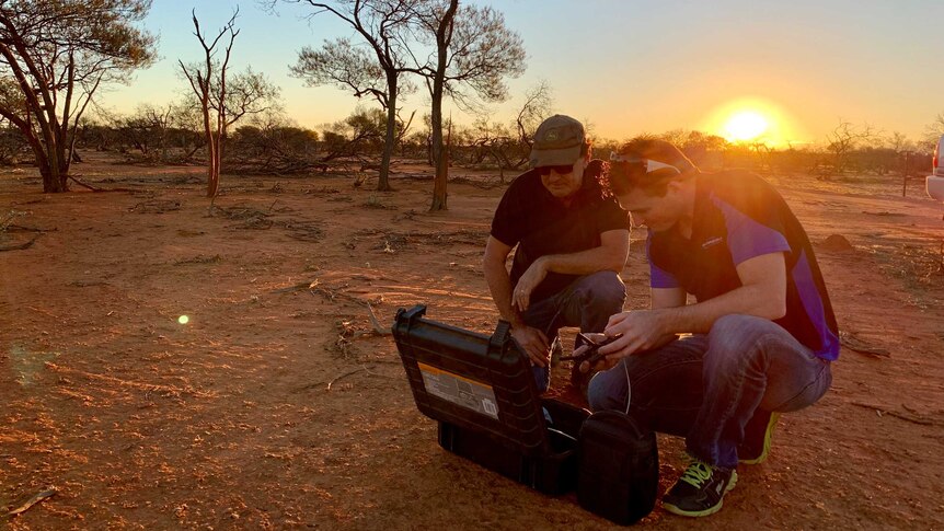 Two men squat over a drone on the ground as the sun goes down behind them