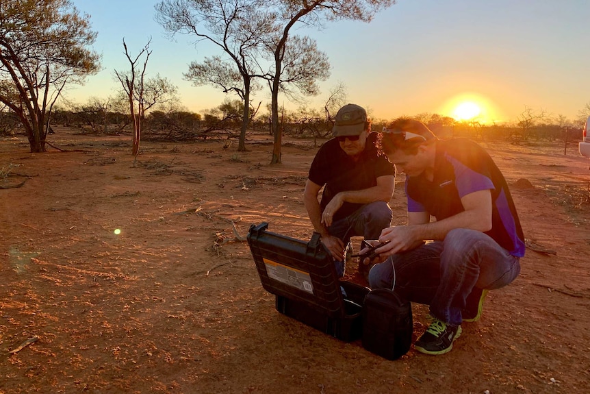 Two men squat over a drone lying on the ground as the sun goes down