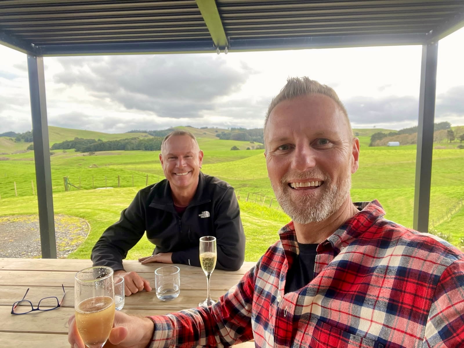A man in a red patterned shirt and a man in a black jacket smile at the camera while at a wine tasting