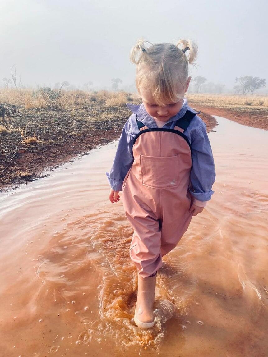 A blonde girl wearing pink overalls and a blue shirt walks through a muddy puddle