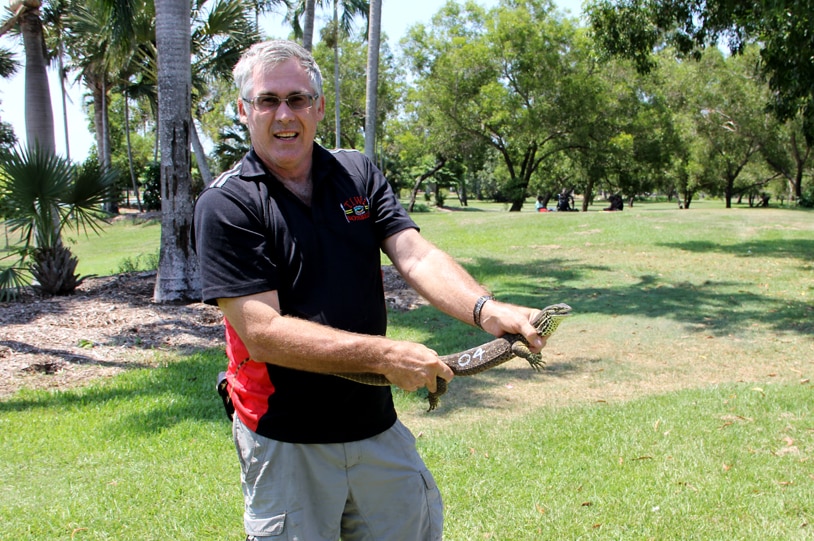 Man standing with small goanna in his hands, smiling at camera.