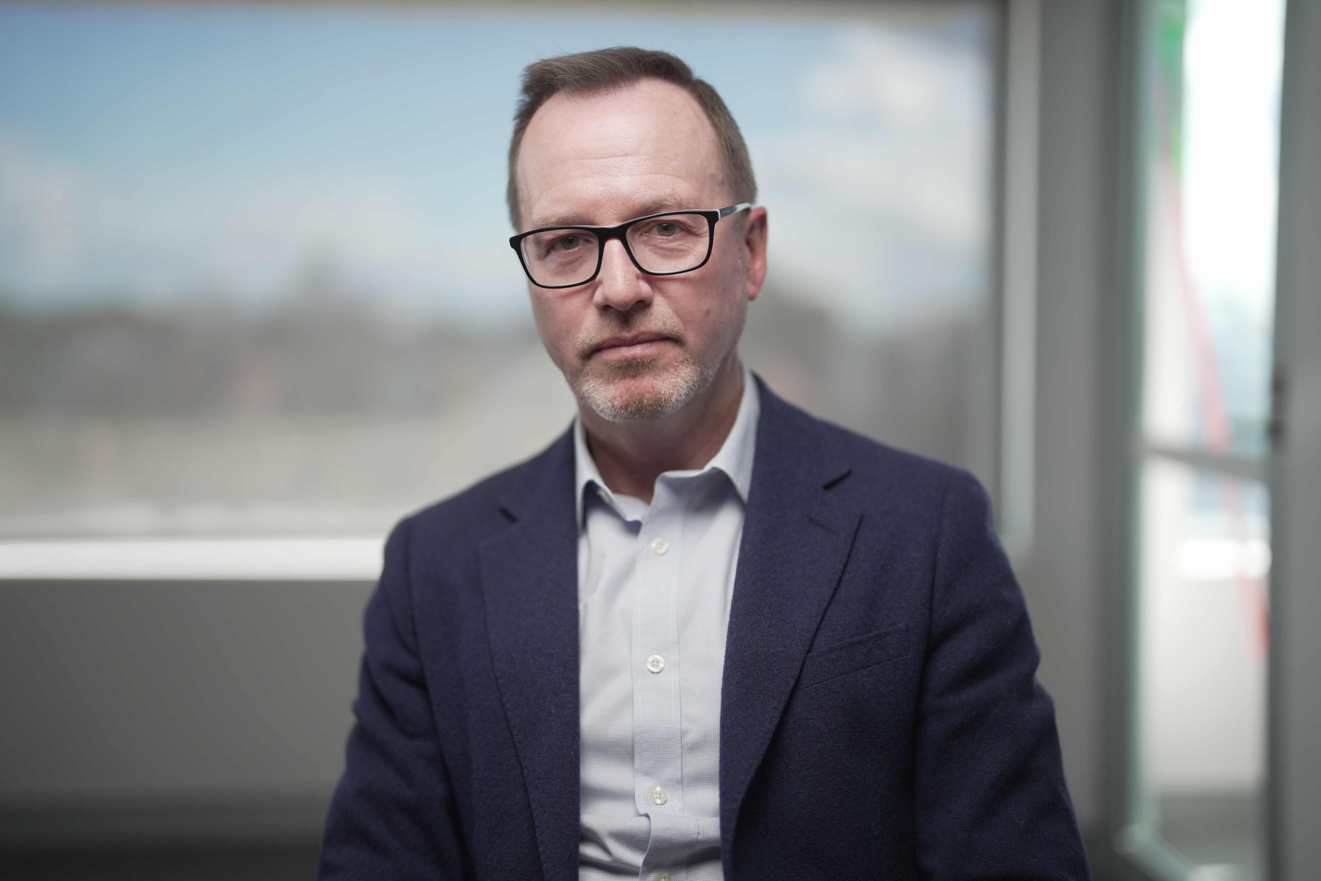 David Shoebridge wears black-rimmed glasses and a navy suit blazer near a window.