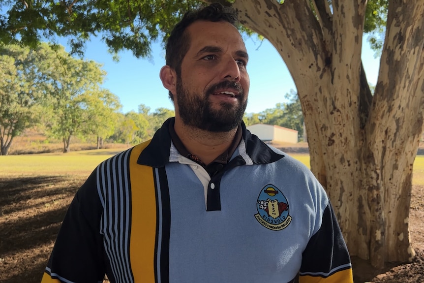 A man in a blue and yellow school jersey, dark beard, stands under a tree, sunny ground and blue skies behind, speaks to camera.